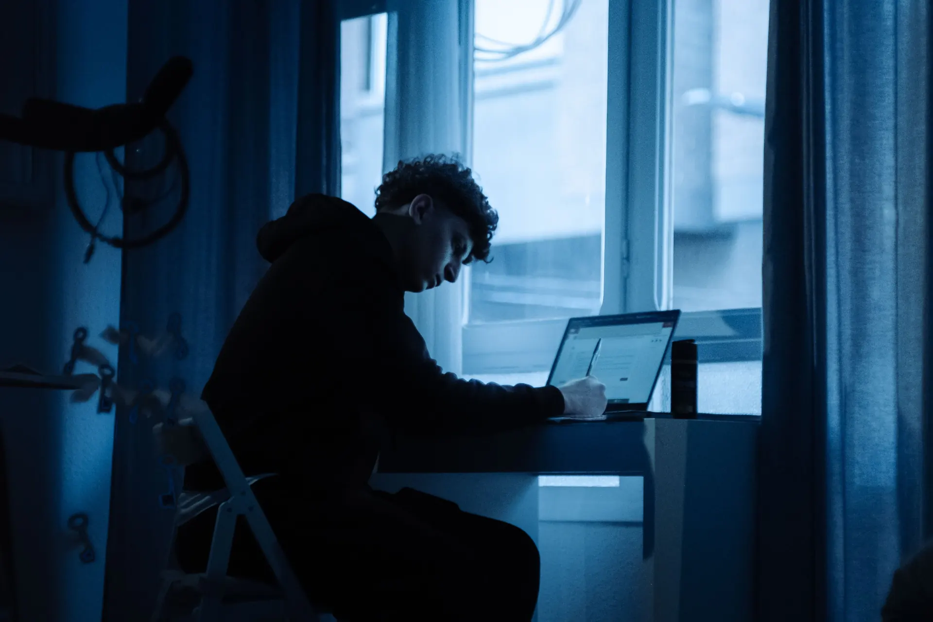 A young man working intently on a laptop beside a window at night